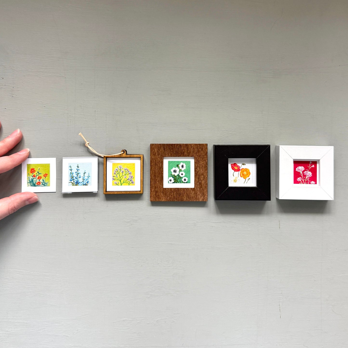 A hand is holding a row of five small framed artworks, each with a different color and style, against a gray wall.