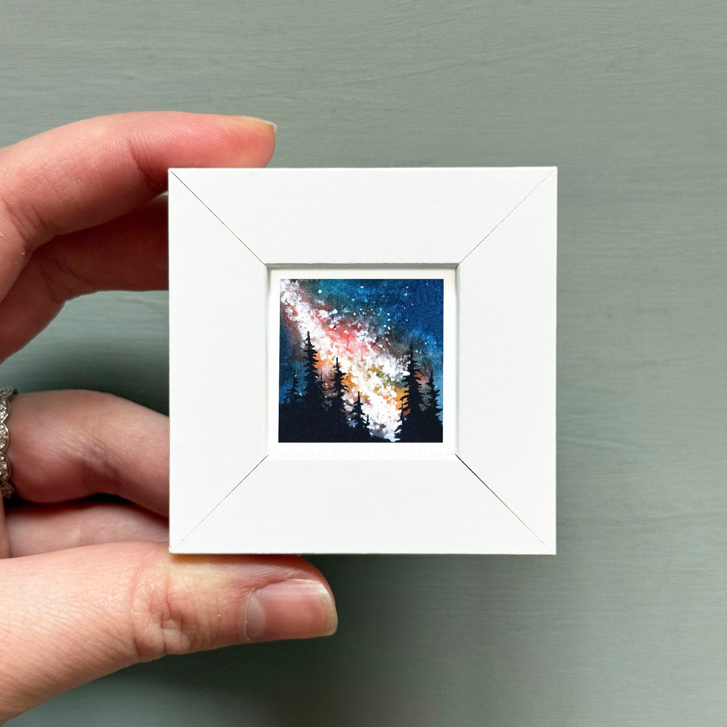 A hand is holding a small white square frame containing a painting of a night sky with stars and silhouettes of trees.