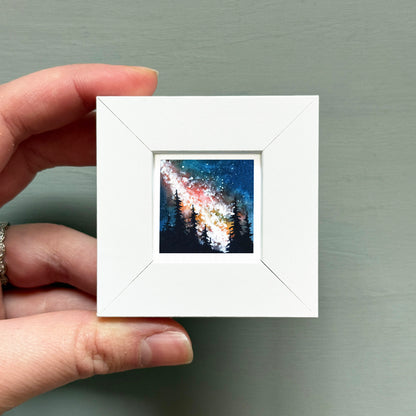 A hand is holding a small white square frame containing a painting of a night sky with stars and silhouettes of trees.