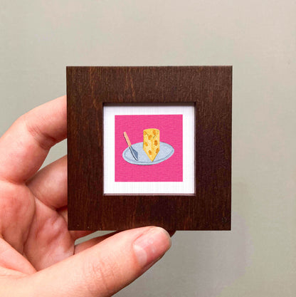 A hand is holding a small square frame containing an illustration of a piece of cheese on a plate.