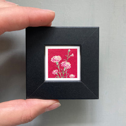 A hand is holding a small black frame containing a vibrant red square with a white border, which displays a close-up of pink flowers.