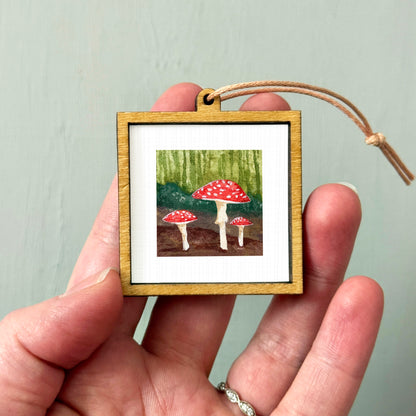 A hand holding a small wooden frame containing a painting of three red mushrooms against a green background.