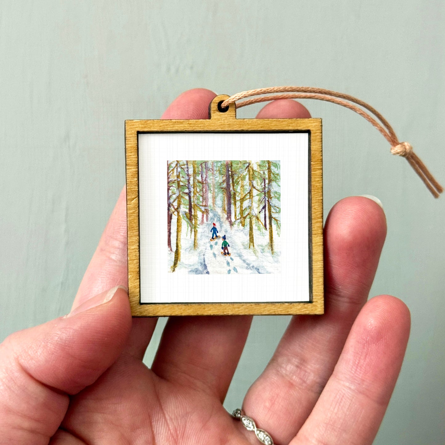 A hand holding a small wooden frame containing a painting of two people skiing in a snowy forest.