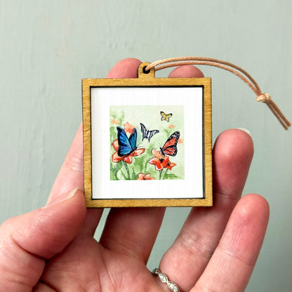 A hand holding a small wooden frame containing a painting of colorful butterflies on flowers.