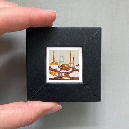 A person's hand is holding a small black picture frame containing an illustration of a table with a roasted turkey, bowls of food, and candles.