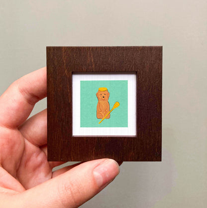 A hand is holding a small wooden frame containing a framed illustration of a bear wearing a yellow hat.