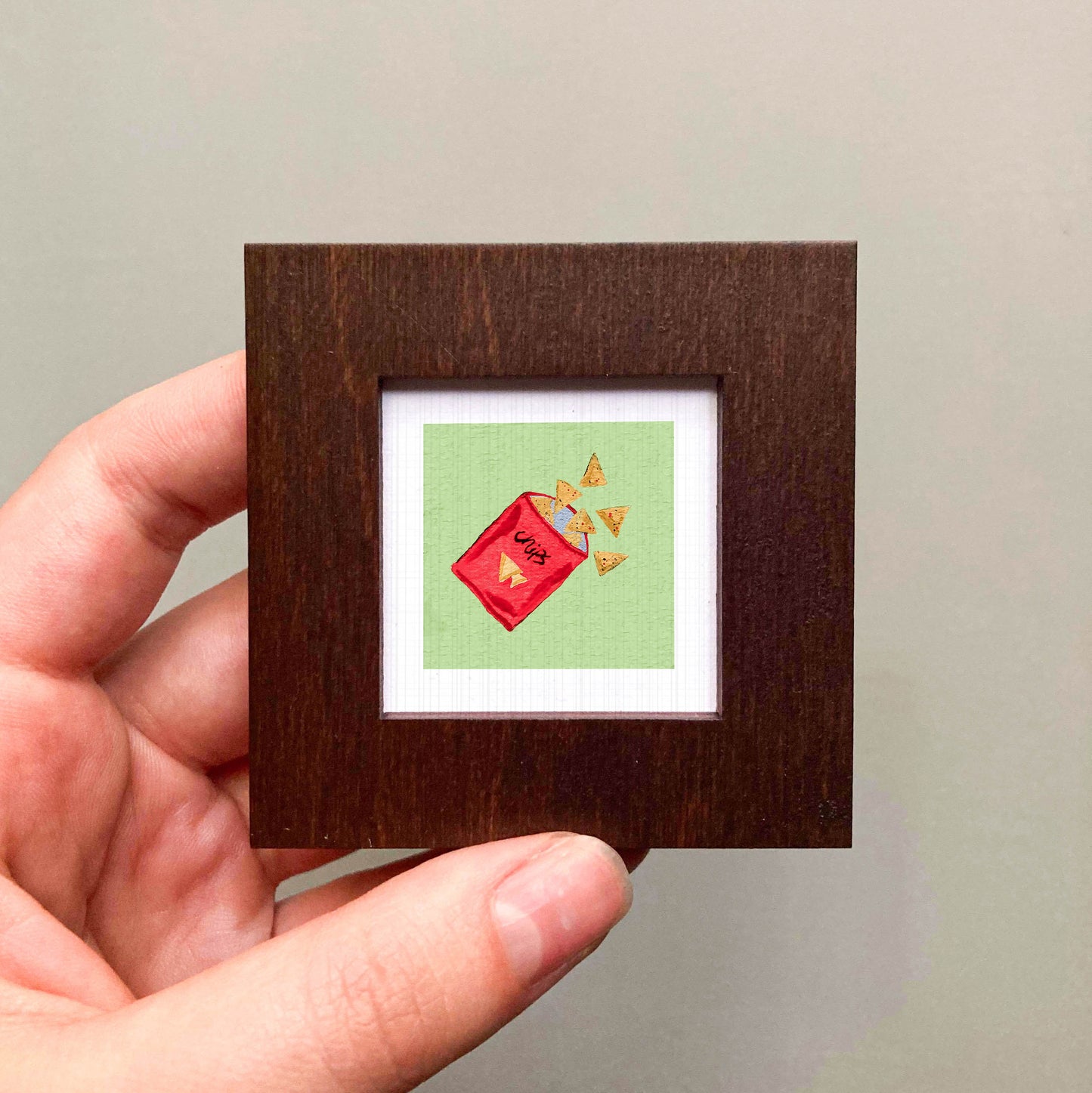 A hand is holding a small wooden frame containing a framed image of a red container with a green background and yellow triangles.