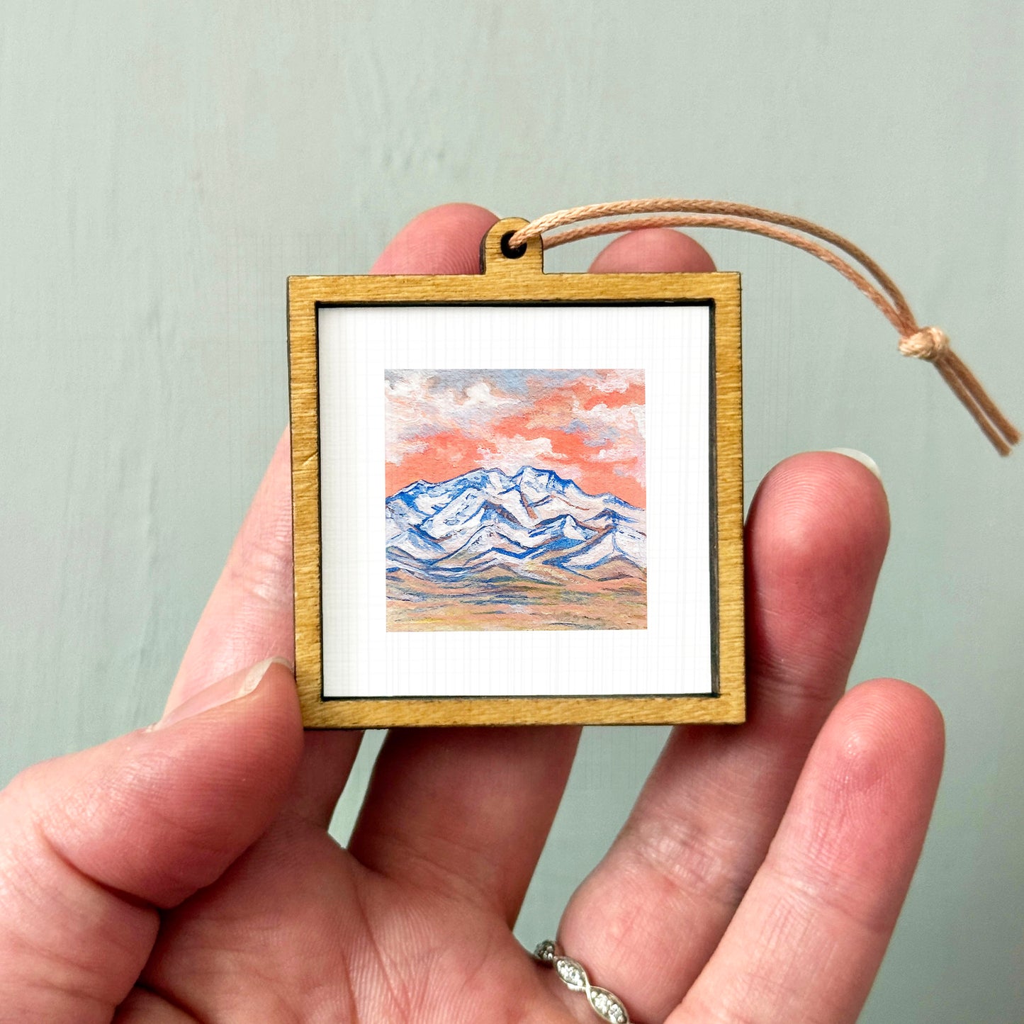 A hand holding a small wooden frame containing a painting of a mountain landscape.
