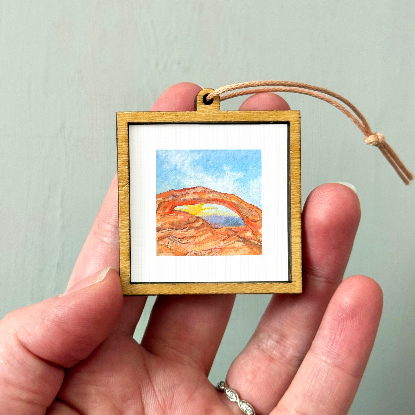 A hand holding a small wooden frame containing a watercolor painting of a desert landscape.