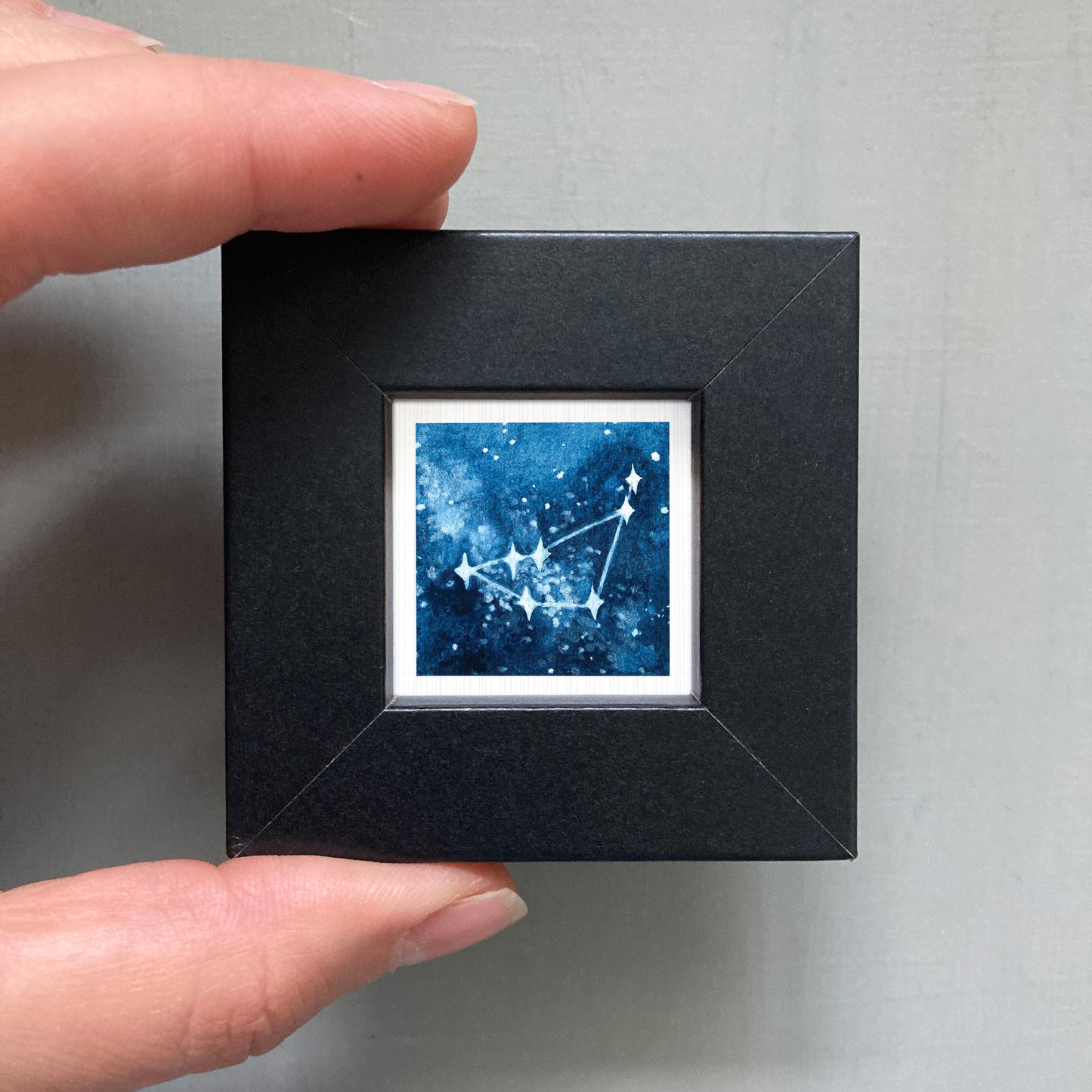 A hand is holding a small black square frame containing a framed image of a starry night sky.
