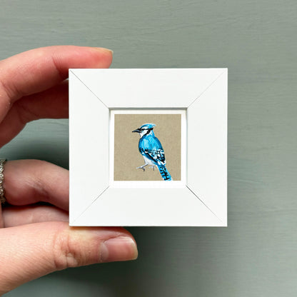 A hand is holding a small white frame containing a painting of a blue jay.
