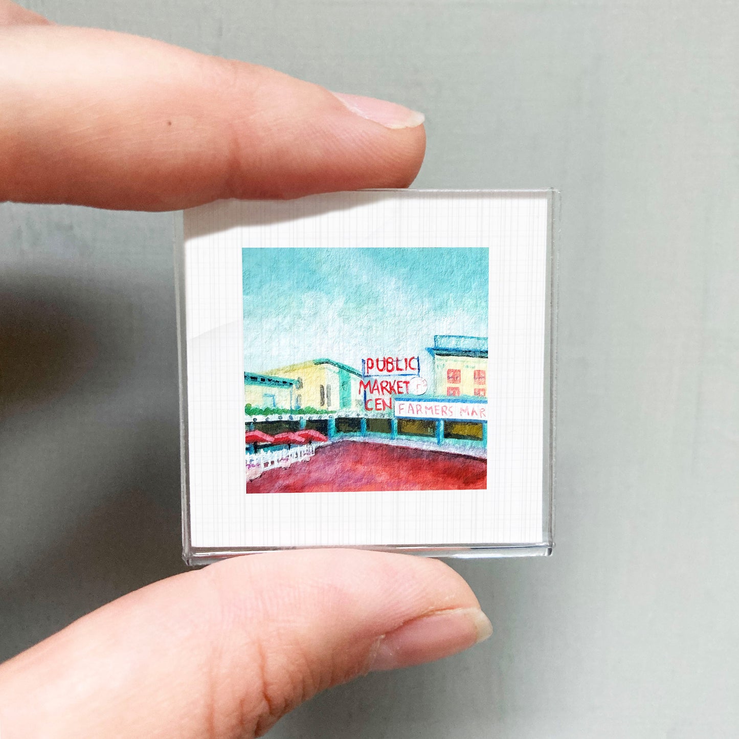 A person's hand holding a small square picture of a colorful street scene with a sign that reads "Public Market".