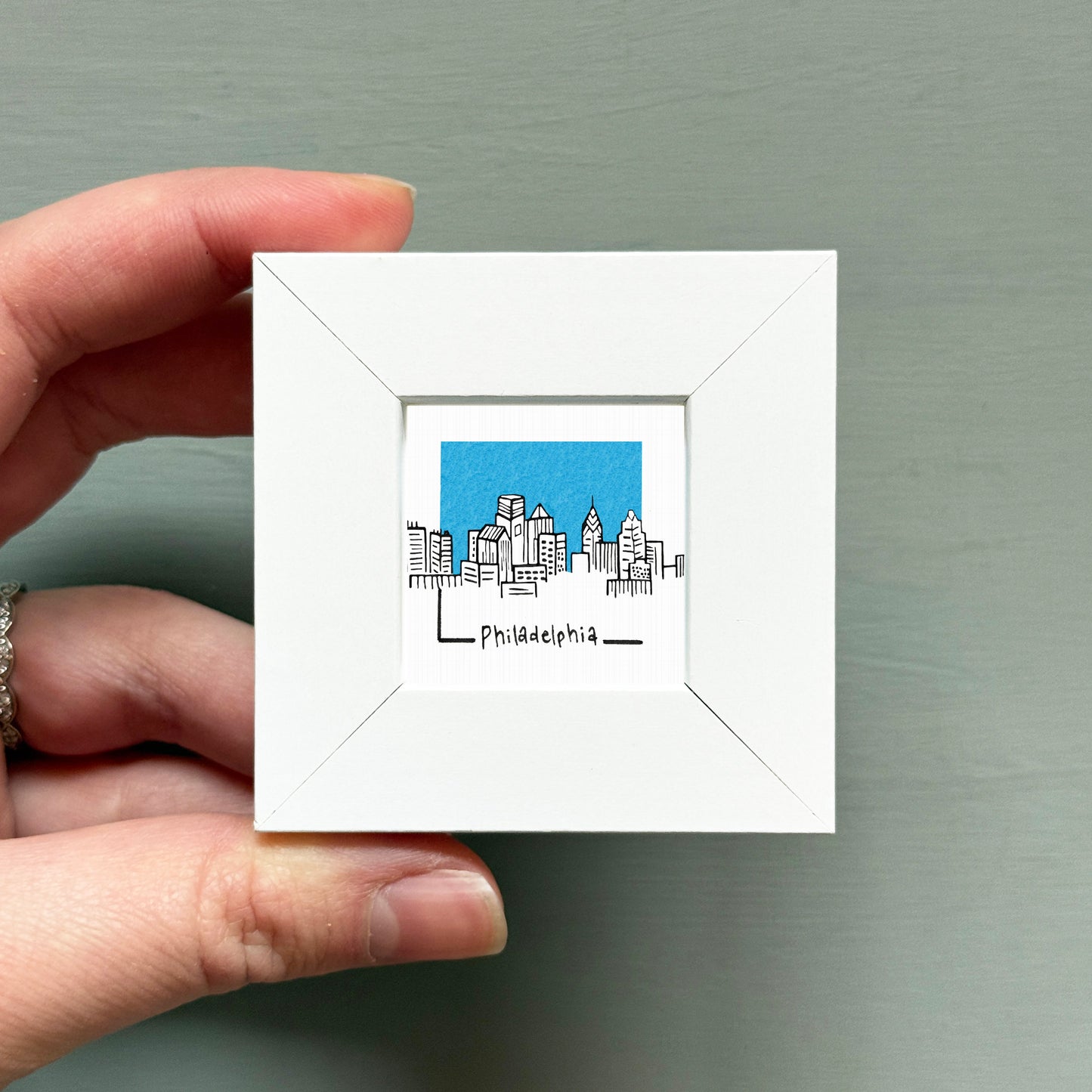 A hand is holding a small white square frame containing a drawing of a city skyline.