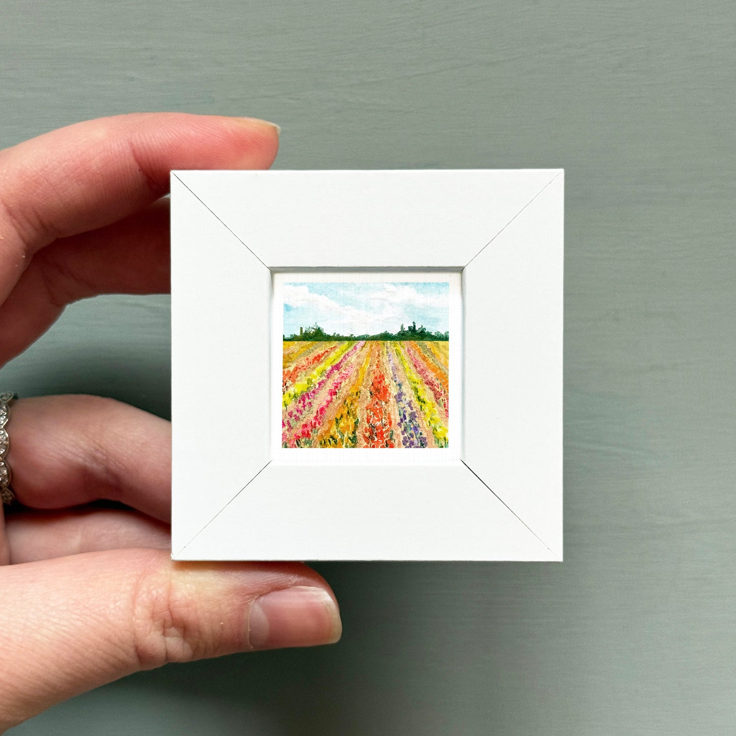 A hand is holding a small white square frame containing a painting of a field of colorful flowers.