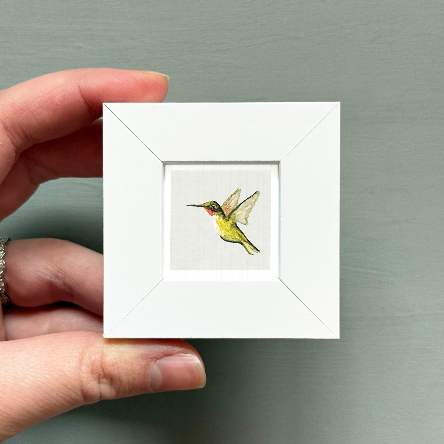 A hand is holding a small white square frame containing a painting of a hummingbird in flight.
