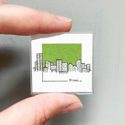 A hand holding a small square magnet with a drawing of a city skyline and the word "Miami" written on it.