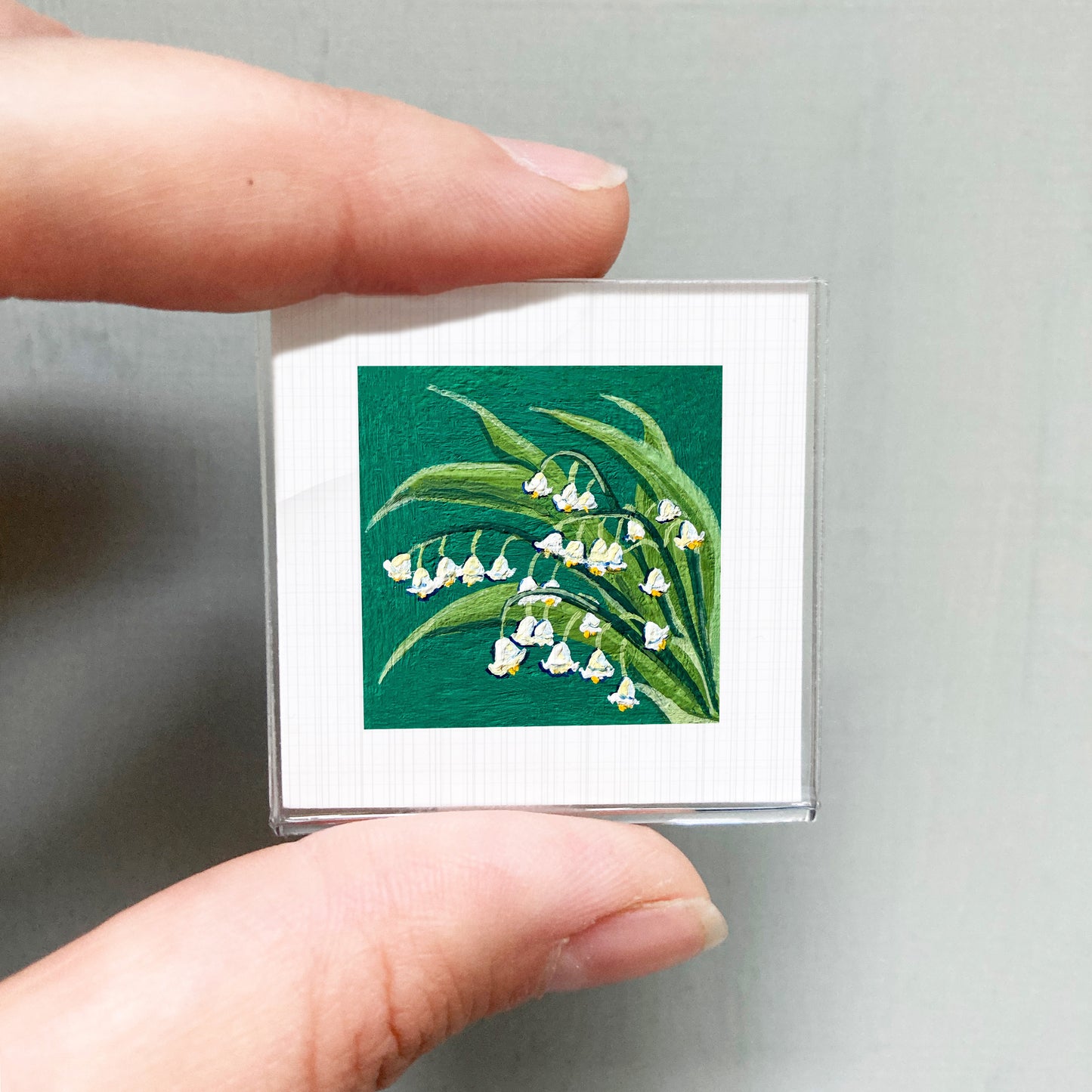 A person's hand holding a small square picture of a lily of the valley plant with white flowers against a green background.