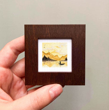 A hand is holding a small, square-shaped wooden frame containing a watercolor painting depicting a person fishing in a lake.