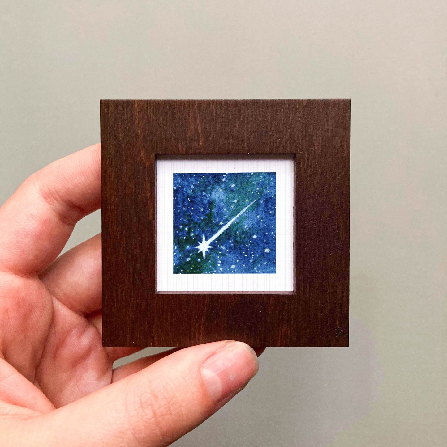 A hand is holding a small wooden frame containing a framed image of a starry night sky.