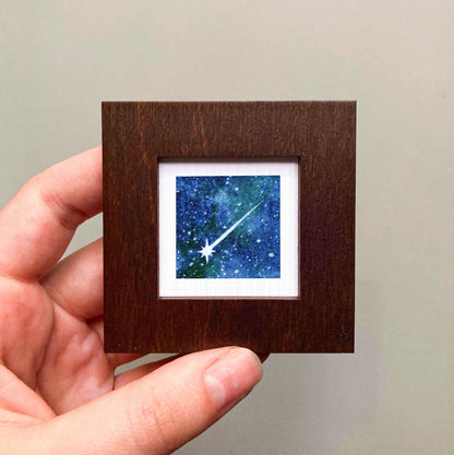 A hand is holding a small wooden frame containing a framed image of a starry night sky.
