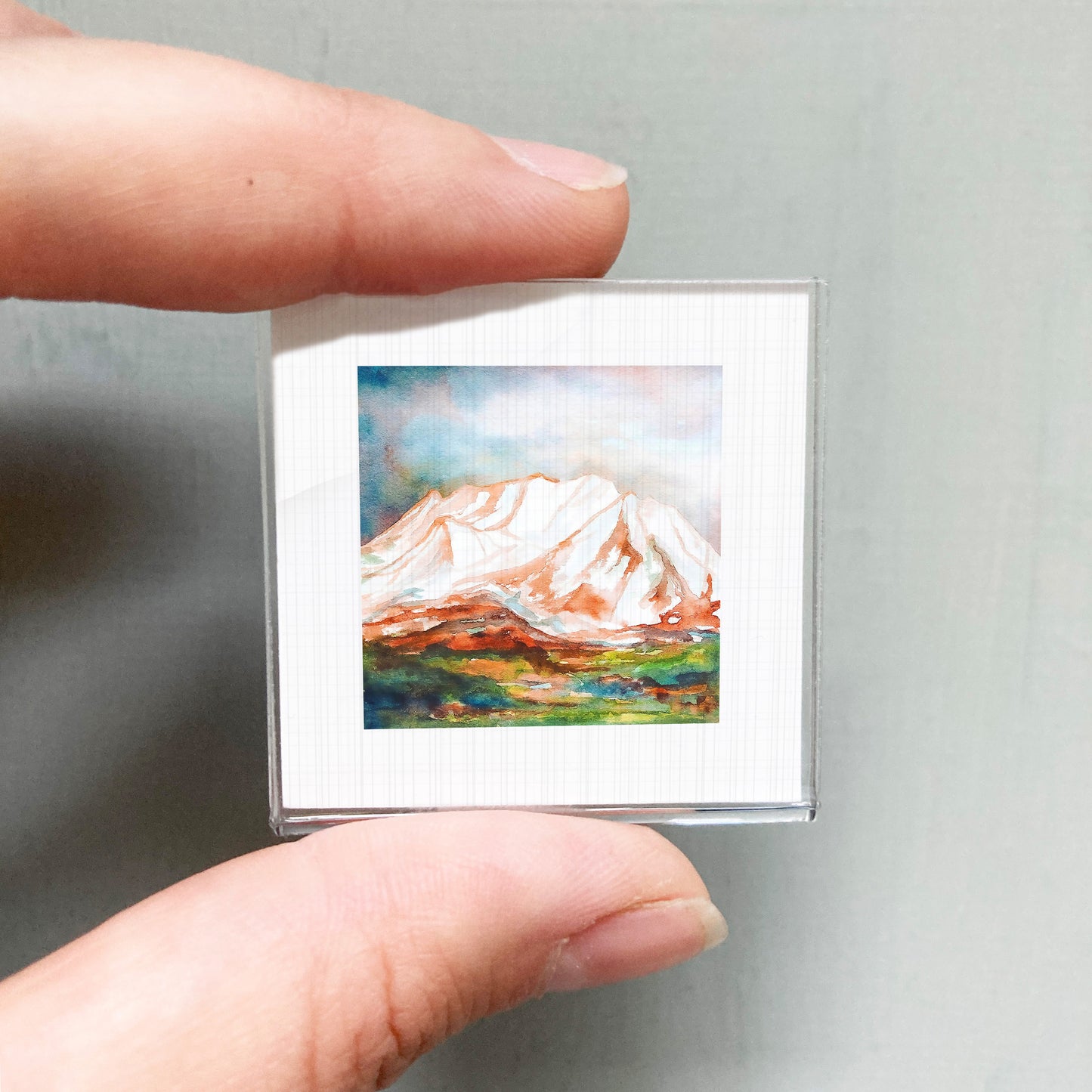 A person's hand holding a small, square-shaped picture of a mountain landscape.