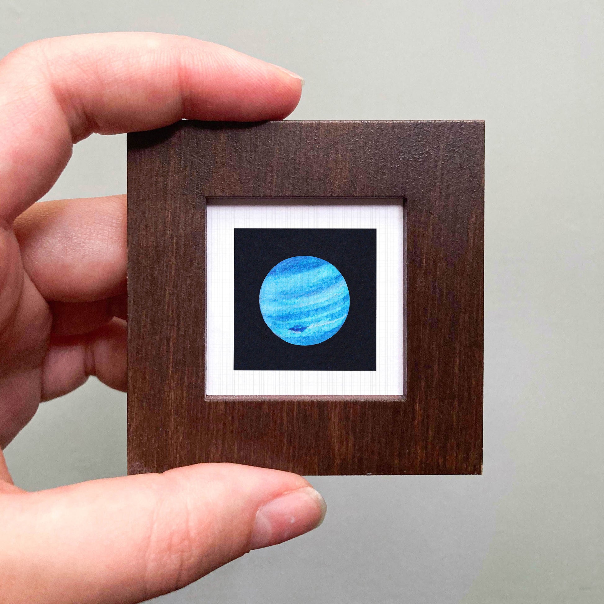 A hand is holding a small wooden frame containing a framed image of a blue planet.