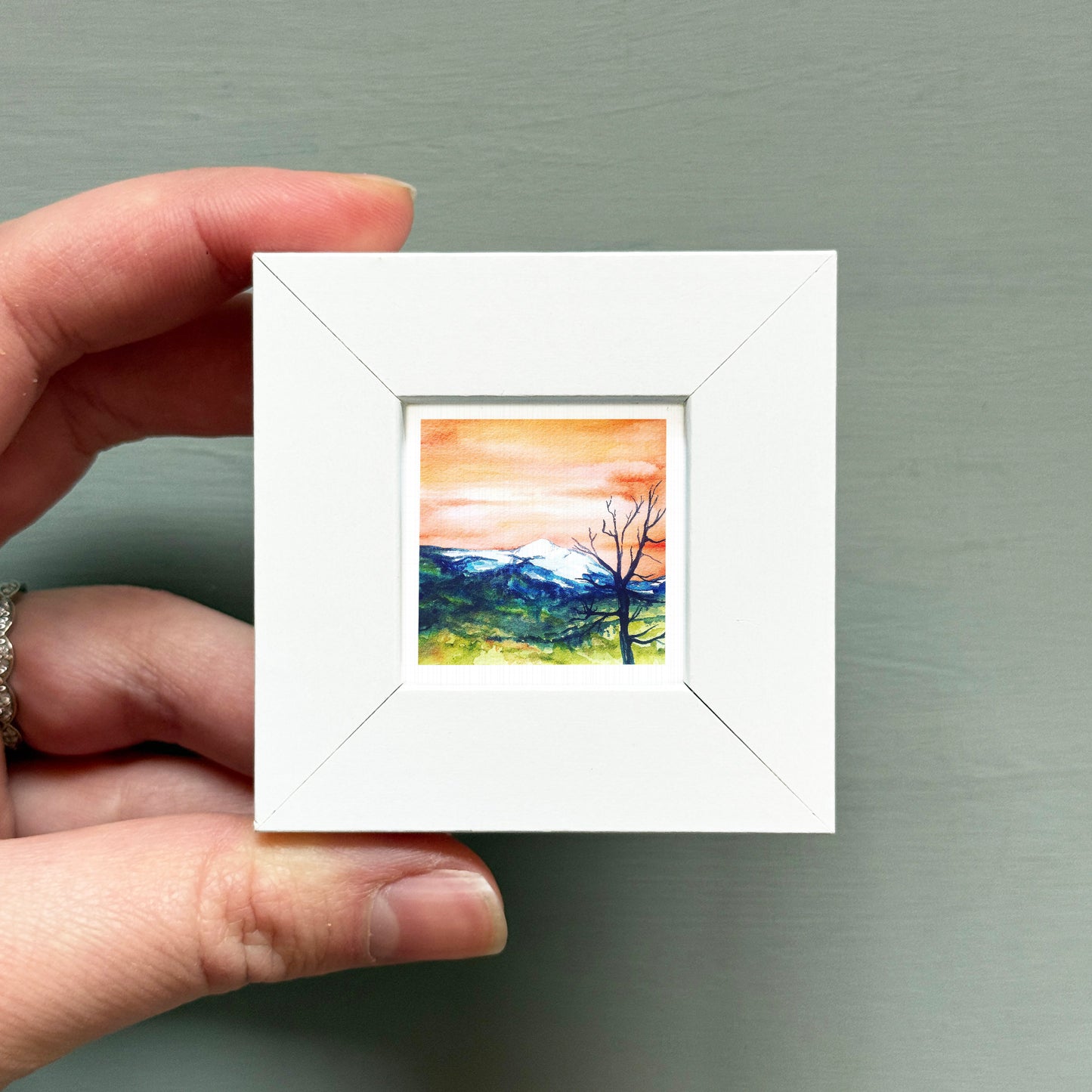A hand is holding a small white square frame containing a watercolor painting of a mountain landscape with a tree in the foreground.