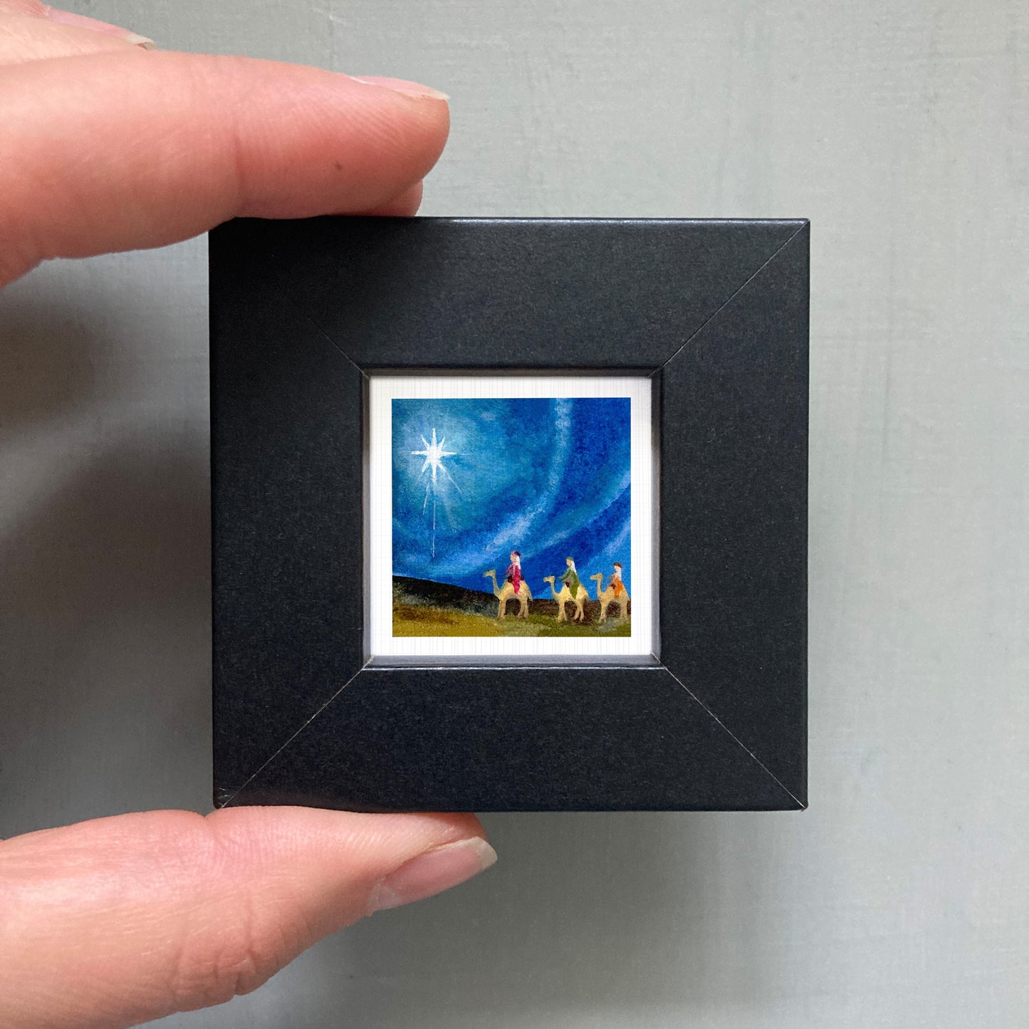 A hand holding a small black frame containing a painting of a group of people on a horse, with a star in the sky.