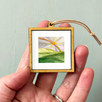 A hand holding a small wooden frame containing a watercolor painting of a landscape with a rainbow in the background.