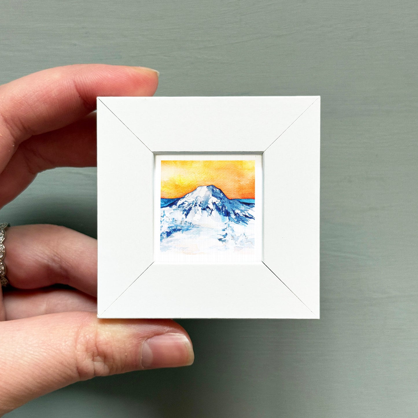 A hand is holding a small white square frame containing a watercolor painting of a mountain landscape.