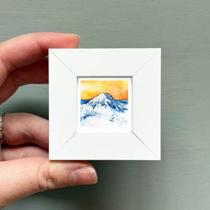 A hand is holding a small white square frame containing a watercolor painting of a mountain landscape.