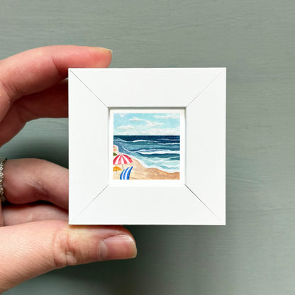 A hand is holding a small white picture frame containing a painting of a beach scene with a blue sky, ocean waves, and a beach umbrella.