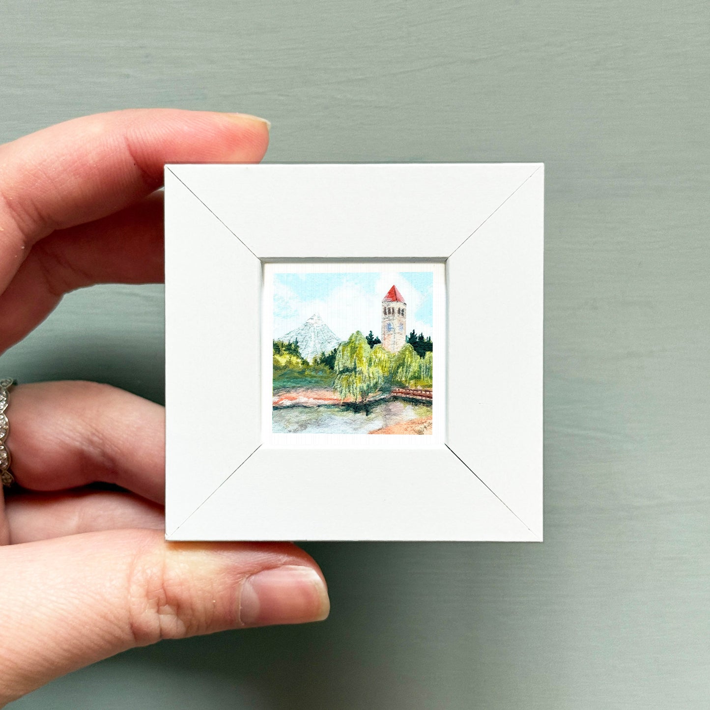 A hand is holding a small white square frame containing a painting of a landscape with a tower in the background.