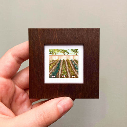 A hand is holding a small, square-shaped picture frame containing a painting of a field with trees and a fence.