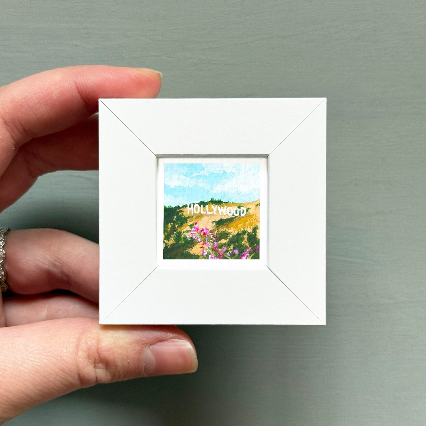 A hand is holding a small white picture frame containing a painting of a scenic landscape with the word "Hollywood" written on it.