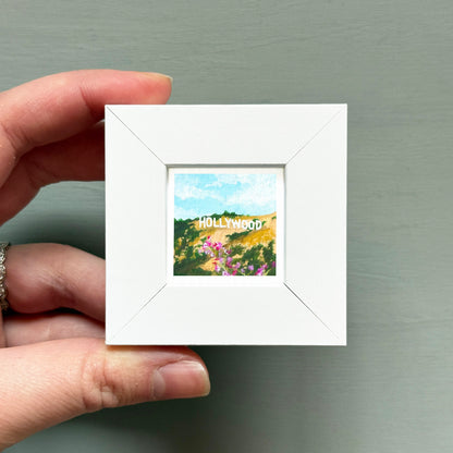 A hand is holding a small white picture frame containing a painting of a scenic landscape with the word "Hollywood" written on it.