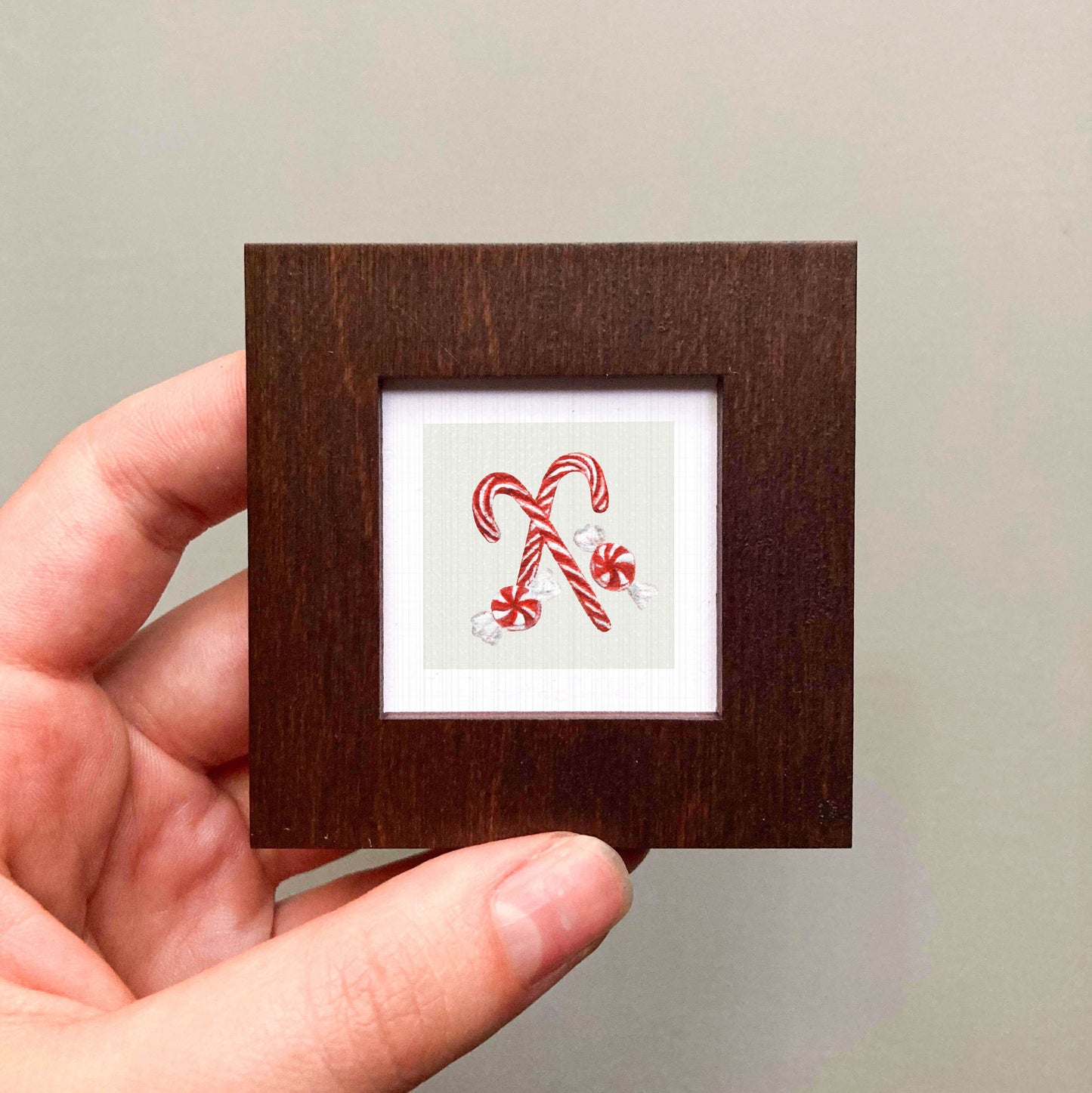 A hand is holding a small wooden frame containing a framed illustration of two red and white striped candy canes.
