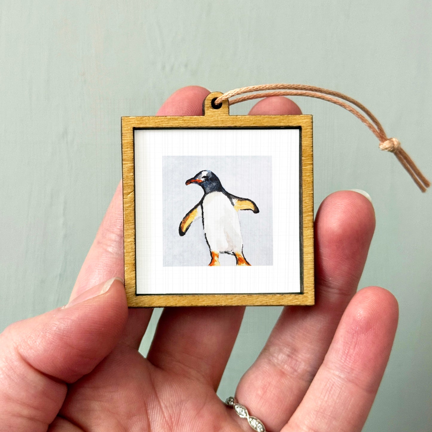 A person's hand holding a small wooden frame containing a picture of a penguin.