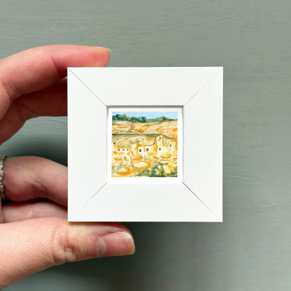 A hand is holding a small white square frame containing a painting of a rural landscape.