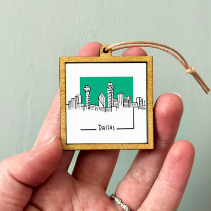 A hand holding a small wooden frame with a picture of the Dallas skyline on it.