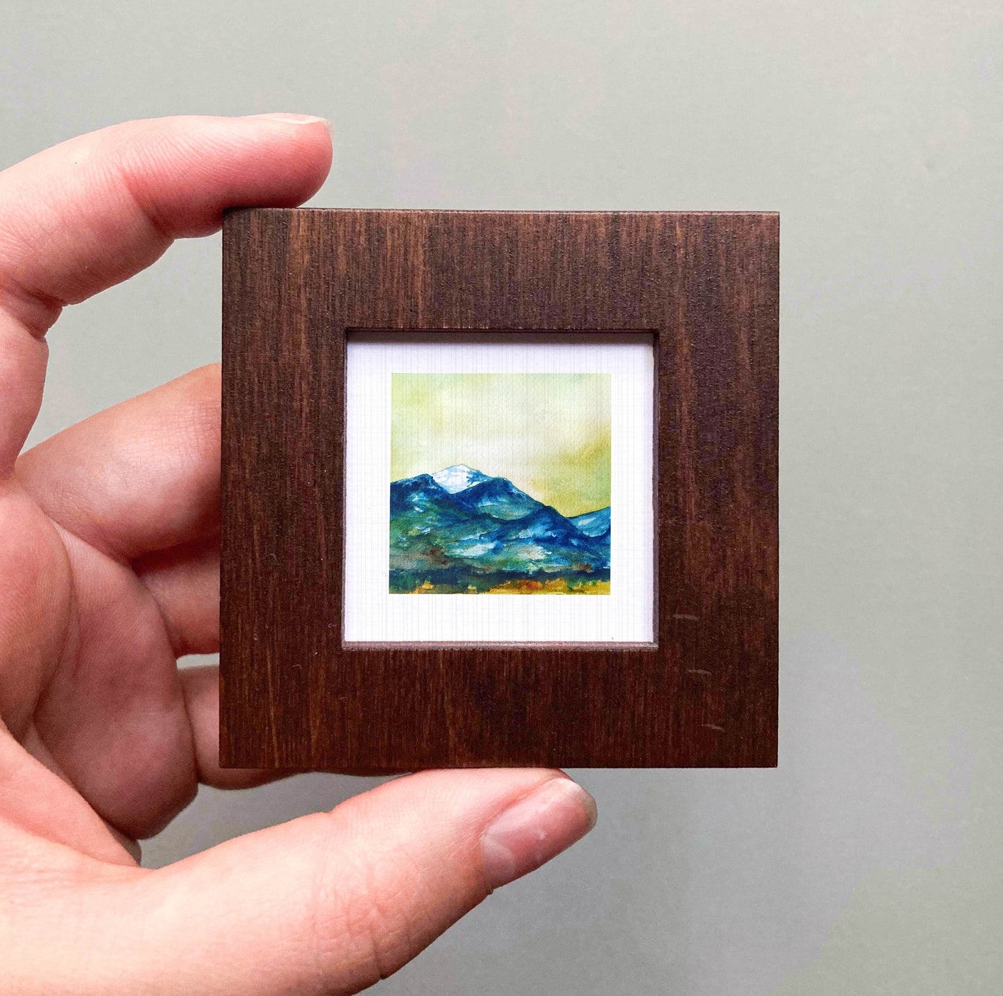 A hand is holding a small wooden frame containing a painting of a mountain landscape.