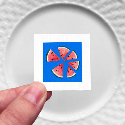 A hand is holding a small square with a vibrant watermelon slice design on a blue background.