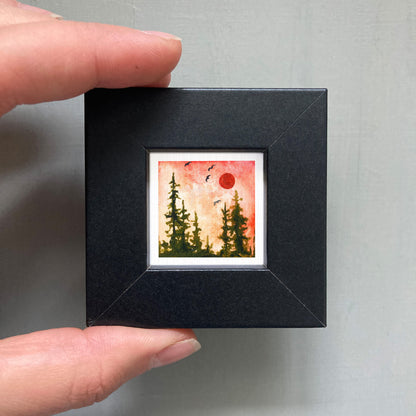 A hand is holding a small black frame containing a framed painting of a forest scene with a red sun in the sky.