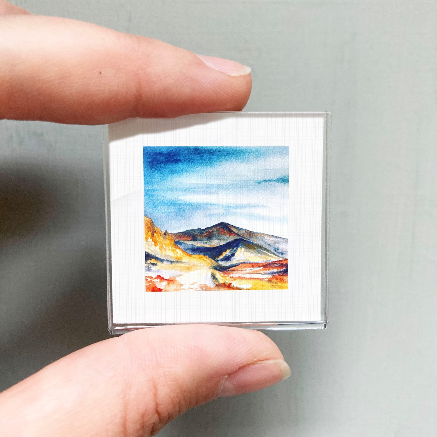 A person's hand is holding a small square picture of a mountain landscape, which appears to be a miniature painting or artwork.