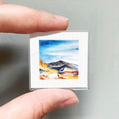A person's hand is holding a small square picture of a mountain landscape, which appears to be a miniature painting or artwork.