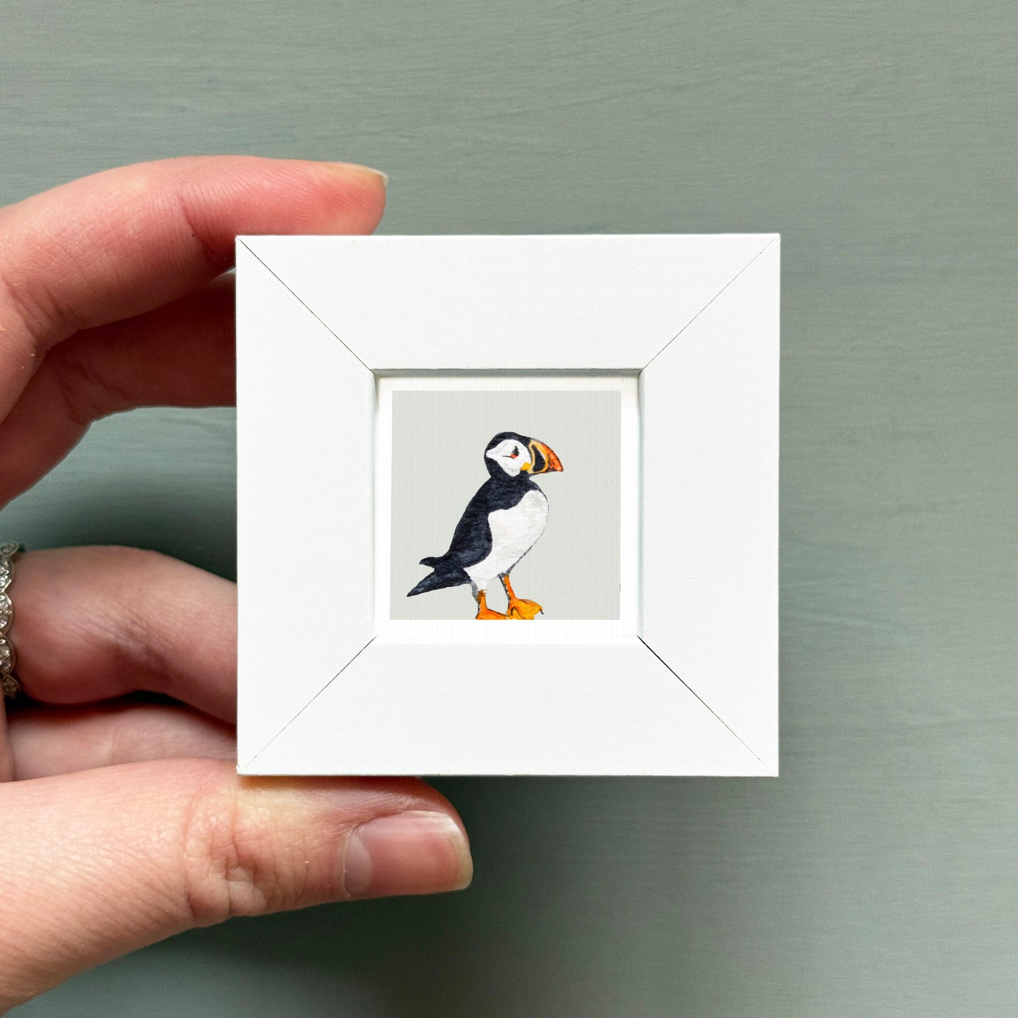 A hand is holding a small white picture frame containing a painting of a black and white bird, which appears to be a puffin.