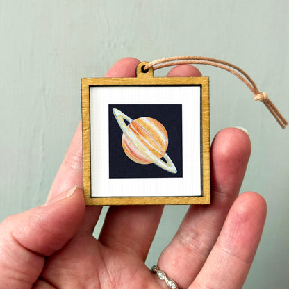 A hand holding a small wooden frame containing a picture of a planet.