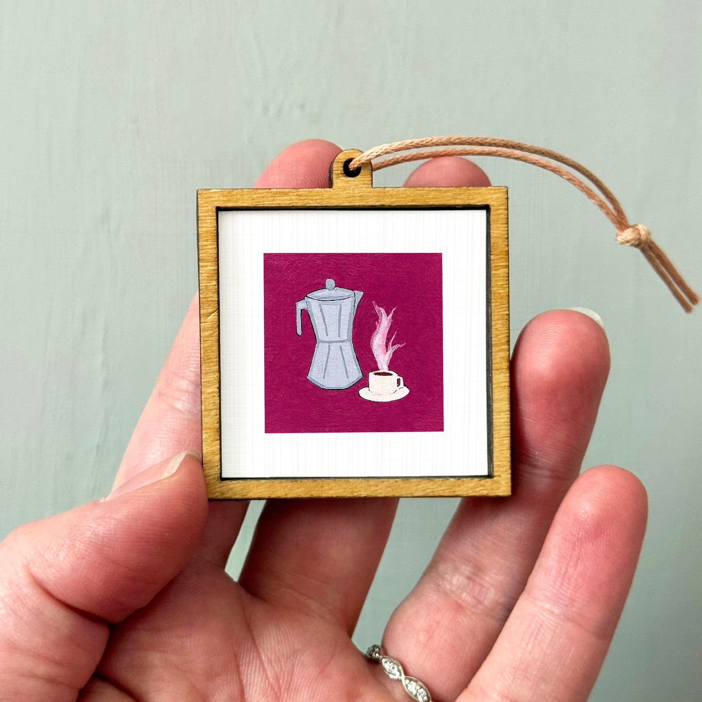 A person's hand holding a small wooden picture frame containing an illustration of a coffee maker and a cup of coffee.