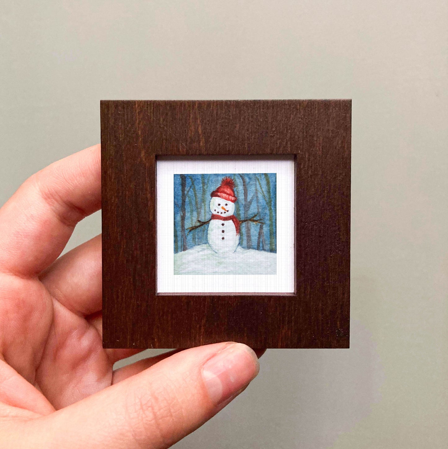 A hand is holding a small, square-shaped picture frame containing a painting of a snowman.