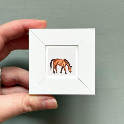 A hand is holding a small white square frame containing a painting of a horse.